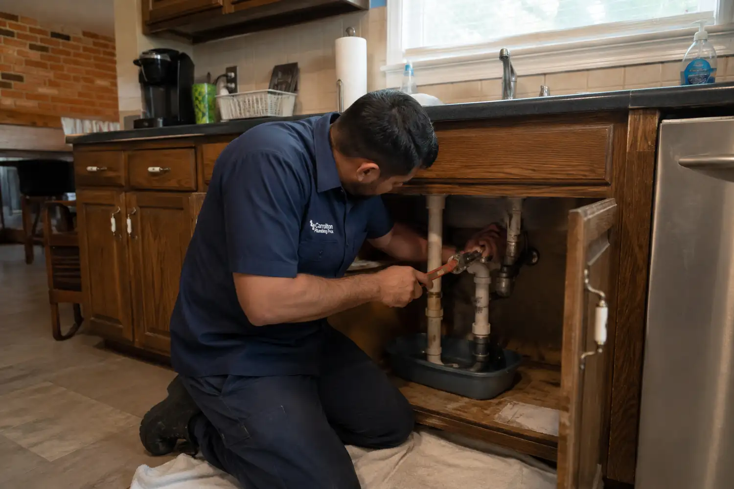 Plumber in Carrollton TX tightening a sink supply connection under a kitchen cabinet, inside a 1980s red brick ranch home